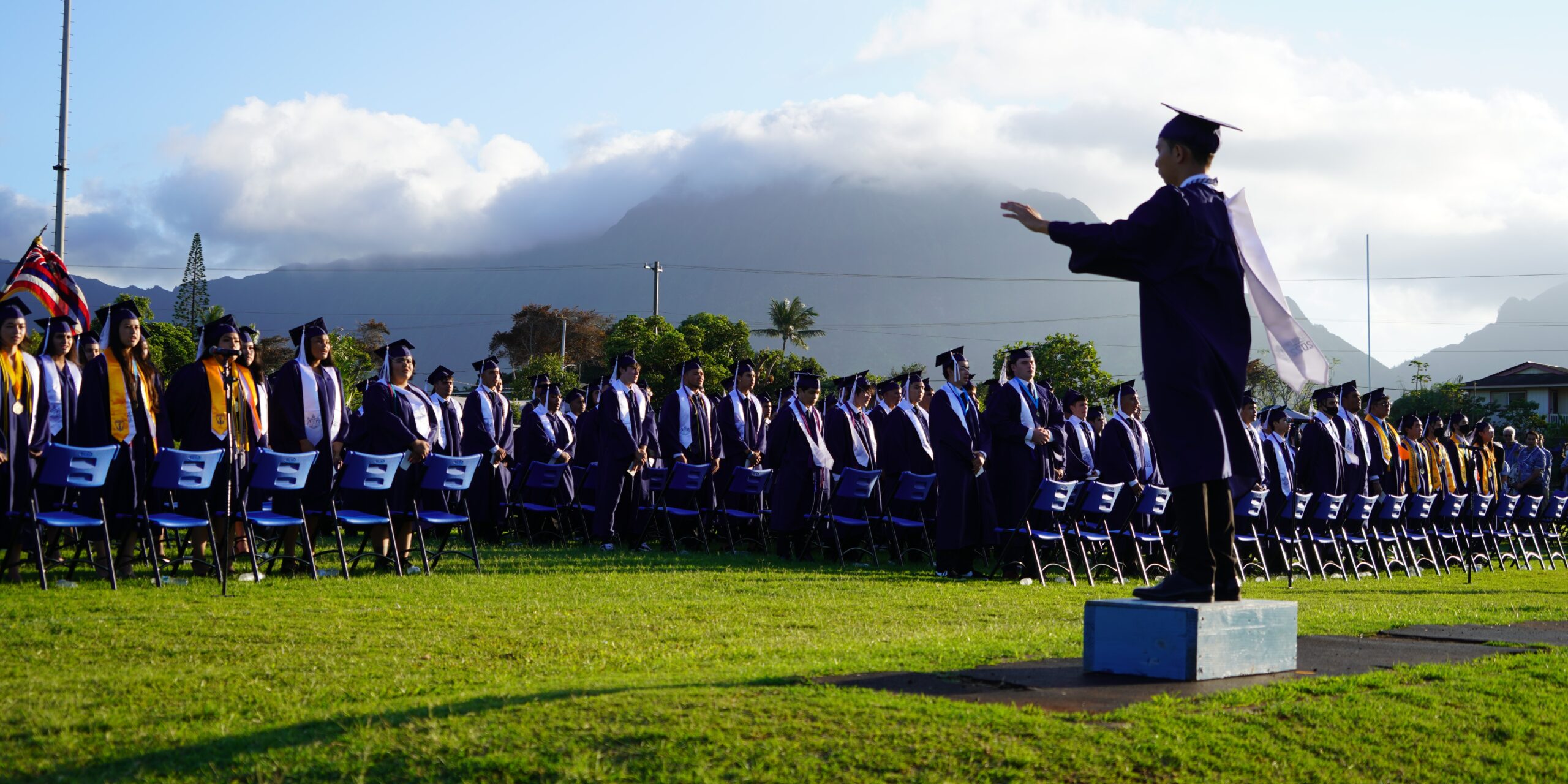 Graduating student standing on top box in a field leading the graduating class in a song.